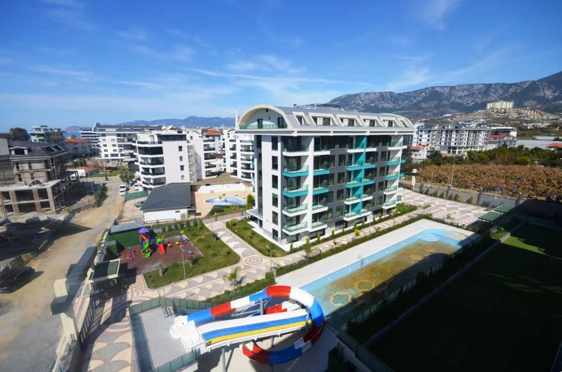 Elevated outdoor view of a luxury residential complex with water slides, a swimming pool, and a playground, as seen in the file DSC_0069 (Orta).JPG, featuring apartments for sale in Alanya.