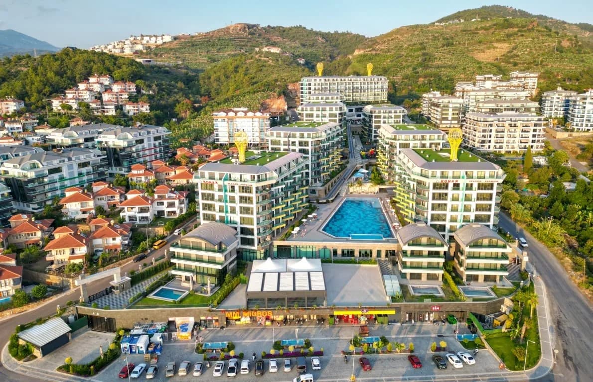 Aerial drone view of Konak Seaside Premium seafront residential complex for sale in Kargıcak, Alanya. Featuring 8 blocks, a large central swimming pool, landscape gardens, and ground-floor commercial shops like Migros and BİM. Taurus Mountains in the background.
