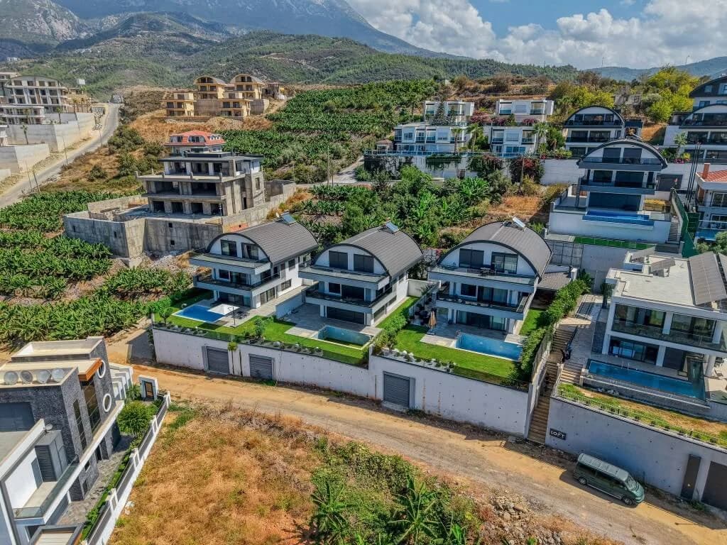 Aerial drone view of the modern 3-story Ada Villas complex, showcasing a luxury villa for sale in Kargicak Alanya with its private swimming pool, garden, and scenic mountain backdrop.